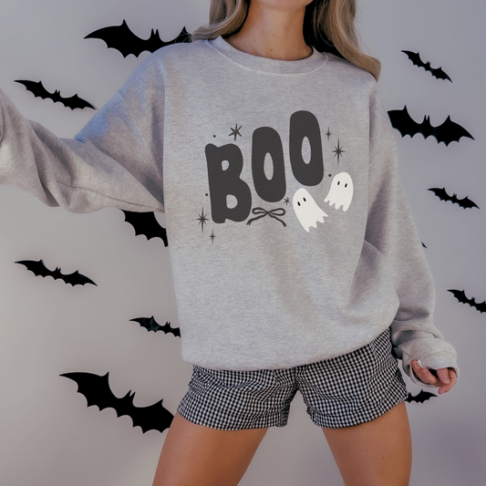 A woman wearing a light heather gray crewneck sweatshirt with a 'BOO' Halloween graphic, paired with black and white gingham shorts, standing against a wall decorated with black bat cutouts.