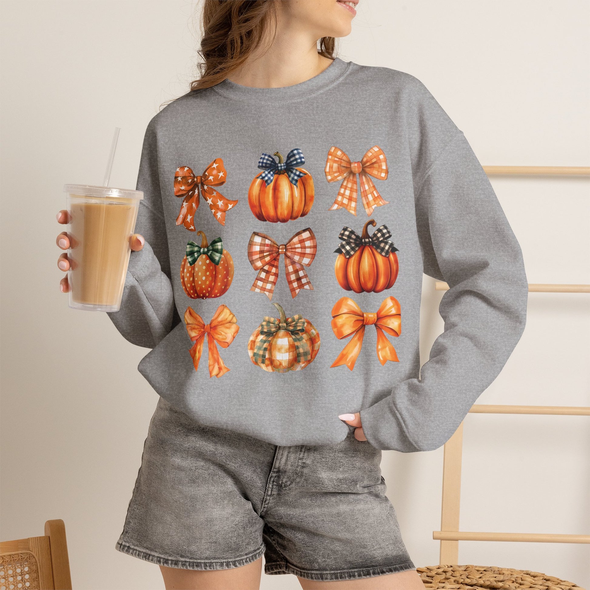 A woman wearing a heather gray crewneck sweatshirt with a graphic print of orange pumpkins and plaid bows, holding an iced coffee in a cafe setting.