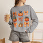 A woman wearing a heather gray crewneck sweatshirt with a graphic print of orange pumpkins and plaid bows, holding an iced coffee in a cafe setting.