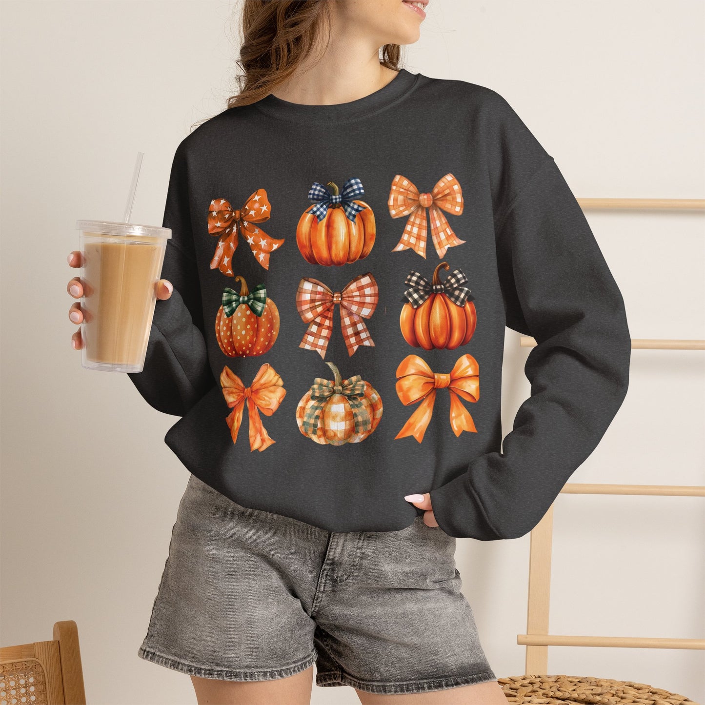 A woman wearing a darker gray crewneck sweatshirt with a graphic print of orange pumpkins and plaid bows, holding an iced coffee in a cafe setting.