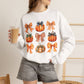 A woman wearing a white crewneck sweatshirt with a graphic print of orange pumpkins and plaid bows, holding a iced coffee in a cafe setting.
