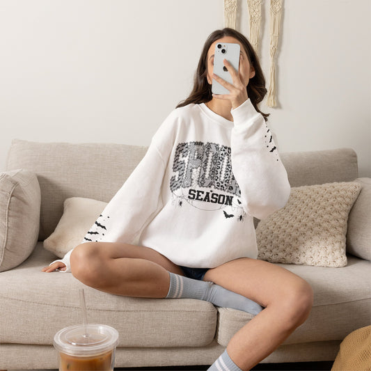 A woman takes a mirror selfie while sitting on a beige couch, wearing a white crewneck sweatshirt with a distressed black graphic that reads 'spooky season'  in a patterned block.