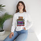 A woman wearing a white crewneck sweatshirt with a colorful graphic of horror movie characters and stacks of VHS tapes, paired with blue jeans, sitting in a bright room next to a potted plant.