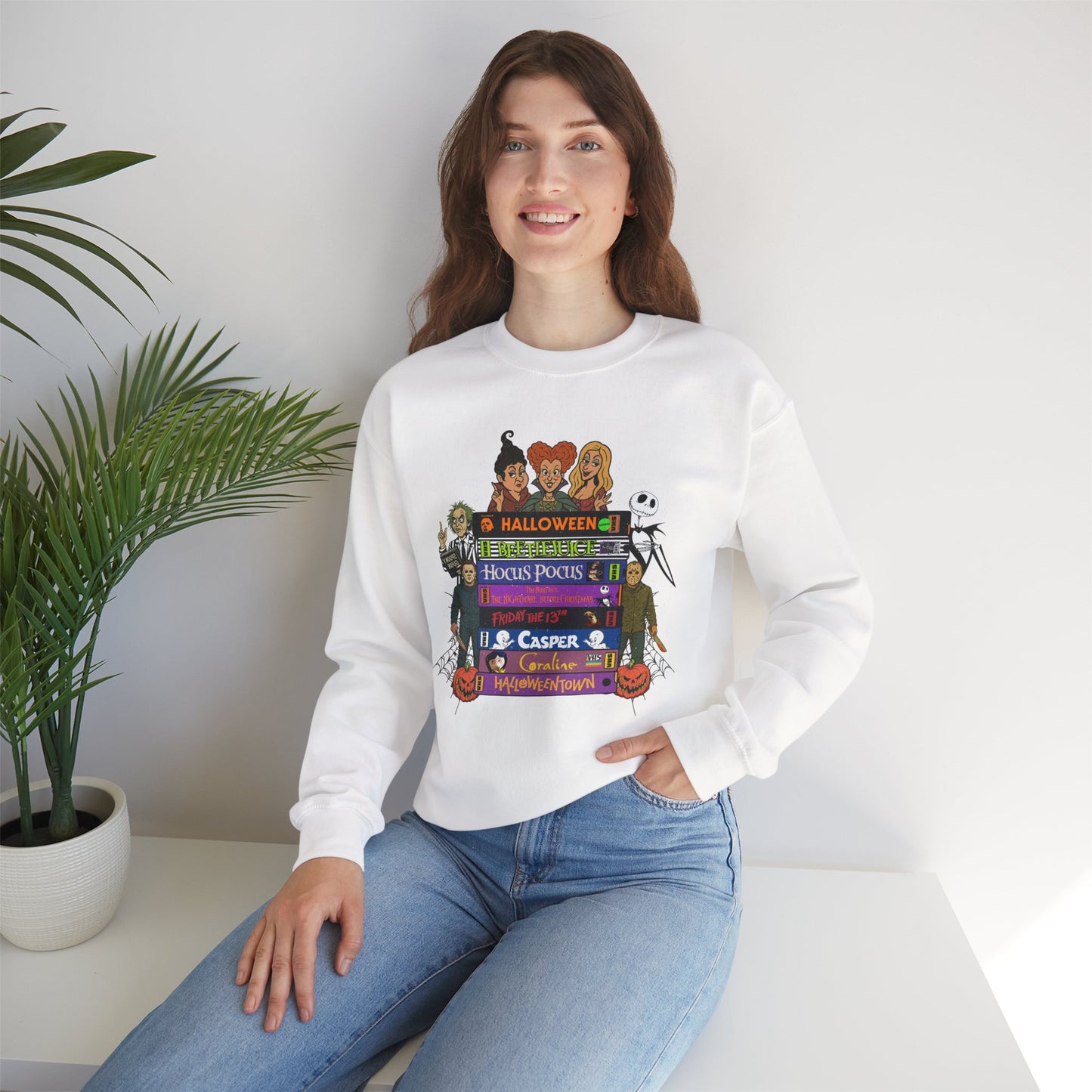 A woman wearing a white crewneck sweatshirt with a colorful graphic of horror movie characters and stacks of VHS tapes, paired with blue jeans, sitting in a bright room next to a potted plant.