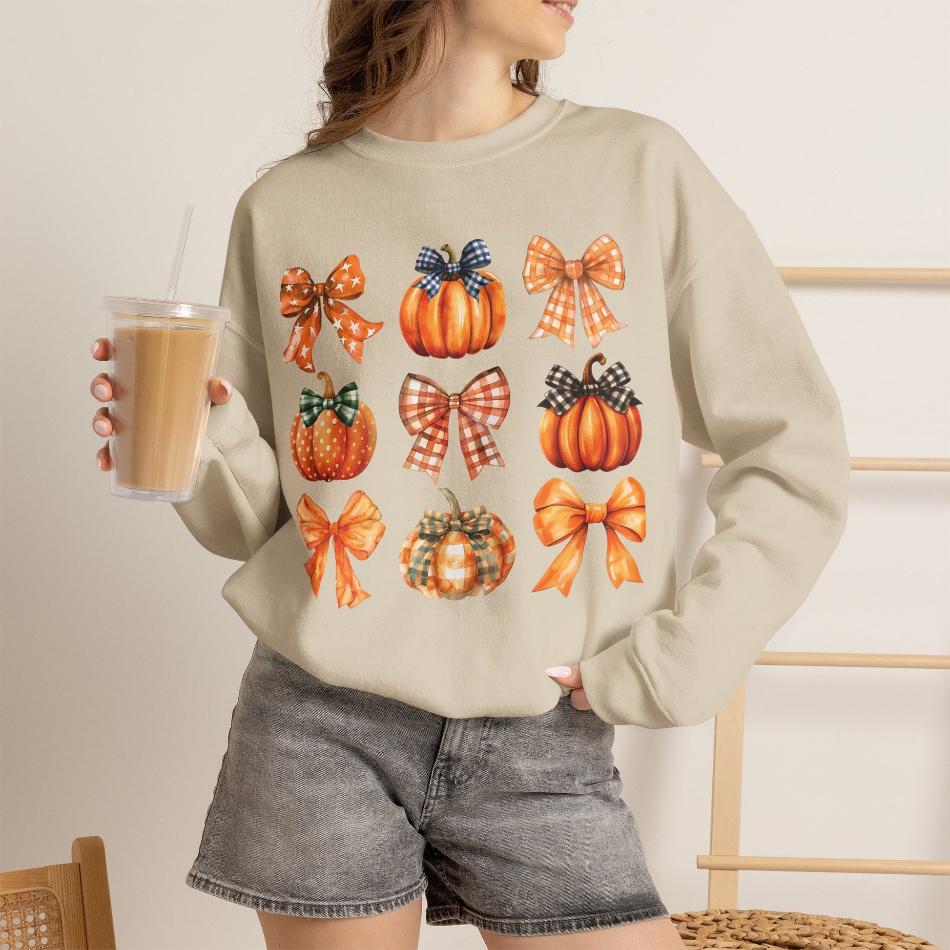 A woman wearing a beige crewneck sweatshirt with a graphic print of orange pumpkins and plaid bows, paired with gray denim shorts and holding a iced coffee.