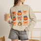 A woman wearing a beige crewneck sweatshirt with a graphic print of orange pumpkins and plaid bows, paired with gray denim shorts and holding a iced coffee.