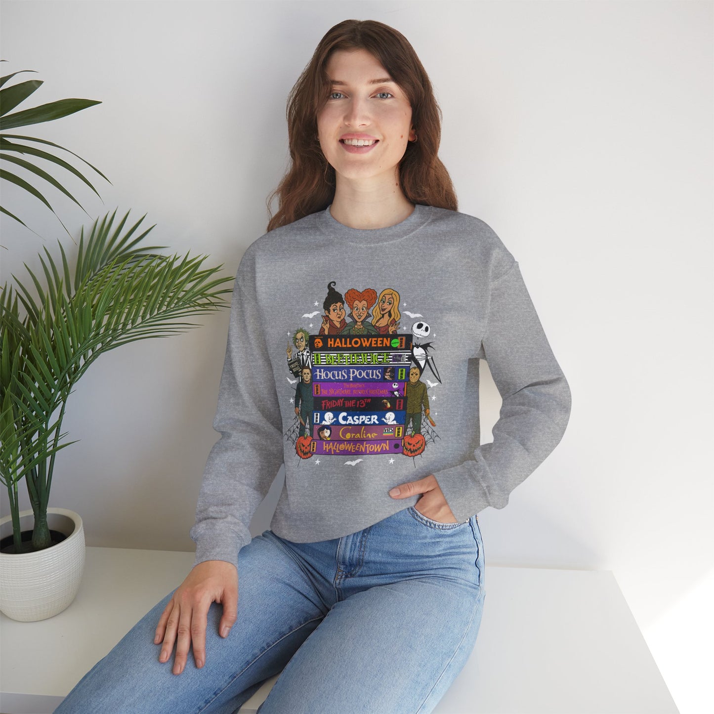 A woman wearing a heather gray crewneck sweatshirt with a colorful Halloween-themed VHS tapes graphic print, paired with blue jeans, sitting in a brightly lit room next to a potted plant.