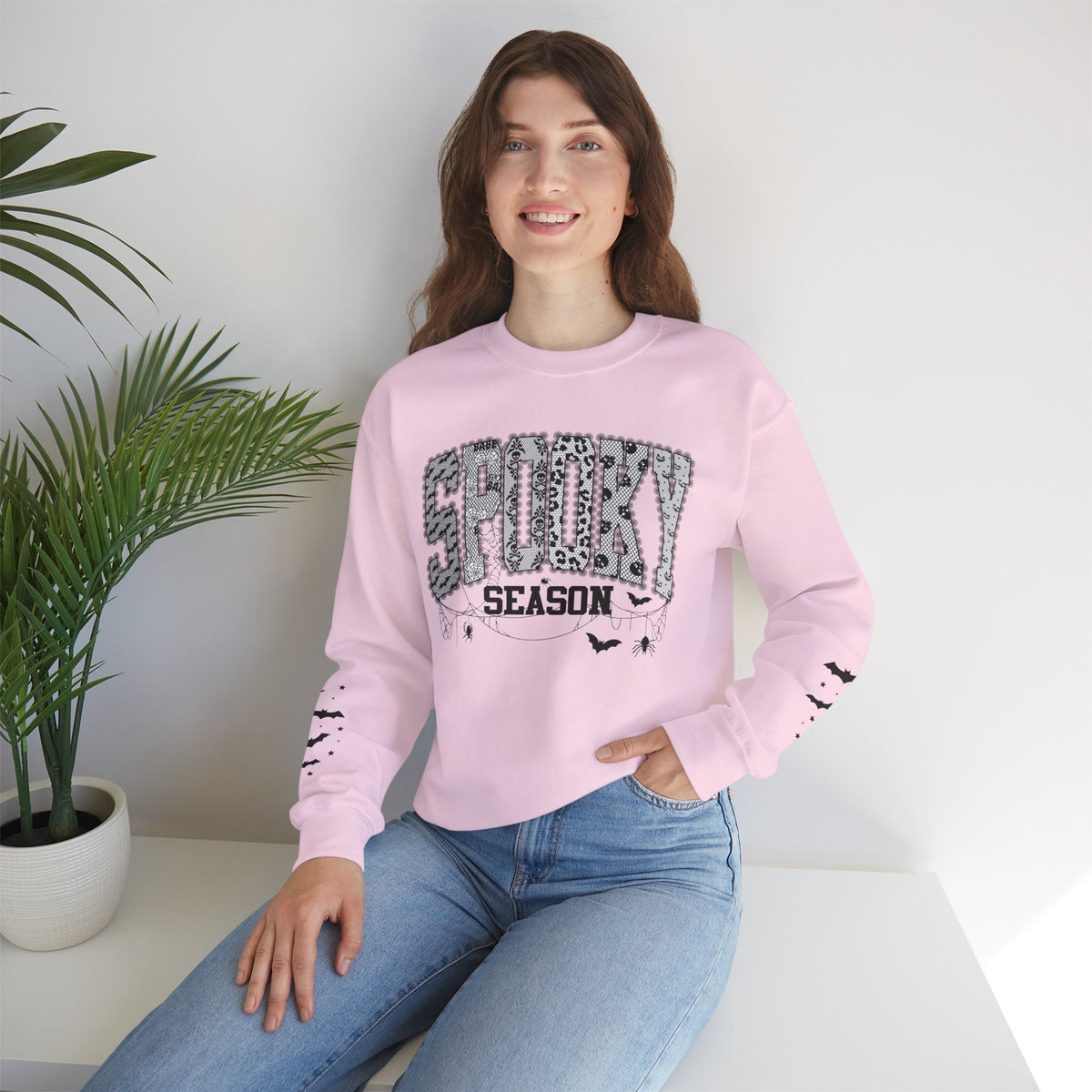 A woman wearing a light pink crewneck sweatshirt with a 'Halloween Season' graphic on the front, paired with blue jeans, sitting in a bright room next to a potted plant.