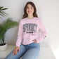 A woman wearing a light pink crewneck sweatshirt with a 'Halloween Season' graphic on the front, paired with blue jeans, sitting in a bright room next to a potted plant.