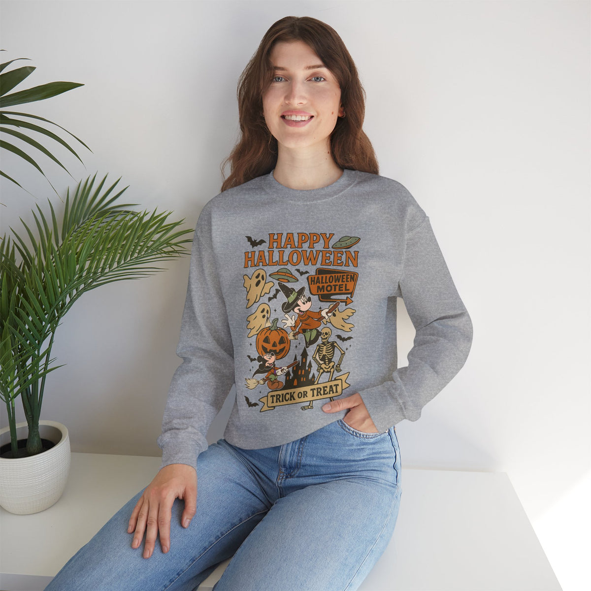A woman wearing a heather gray long-sleeve sweatshirt with a colorful 'Happy Halloween' graphic print, paired with blue jeans, sitting on a white bench next to a potted plant.