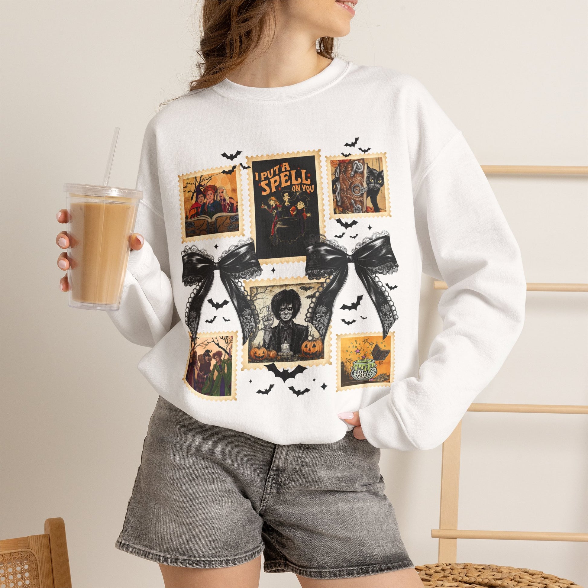 Woman wearing a white crewneck sweatshirt featuring a vintage witch-themed Halloween collage with black bows, bats, pumpkins, and orange accents.
