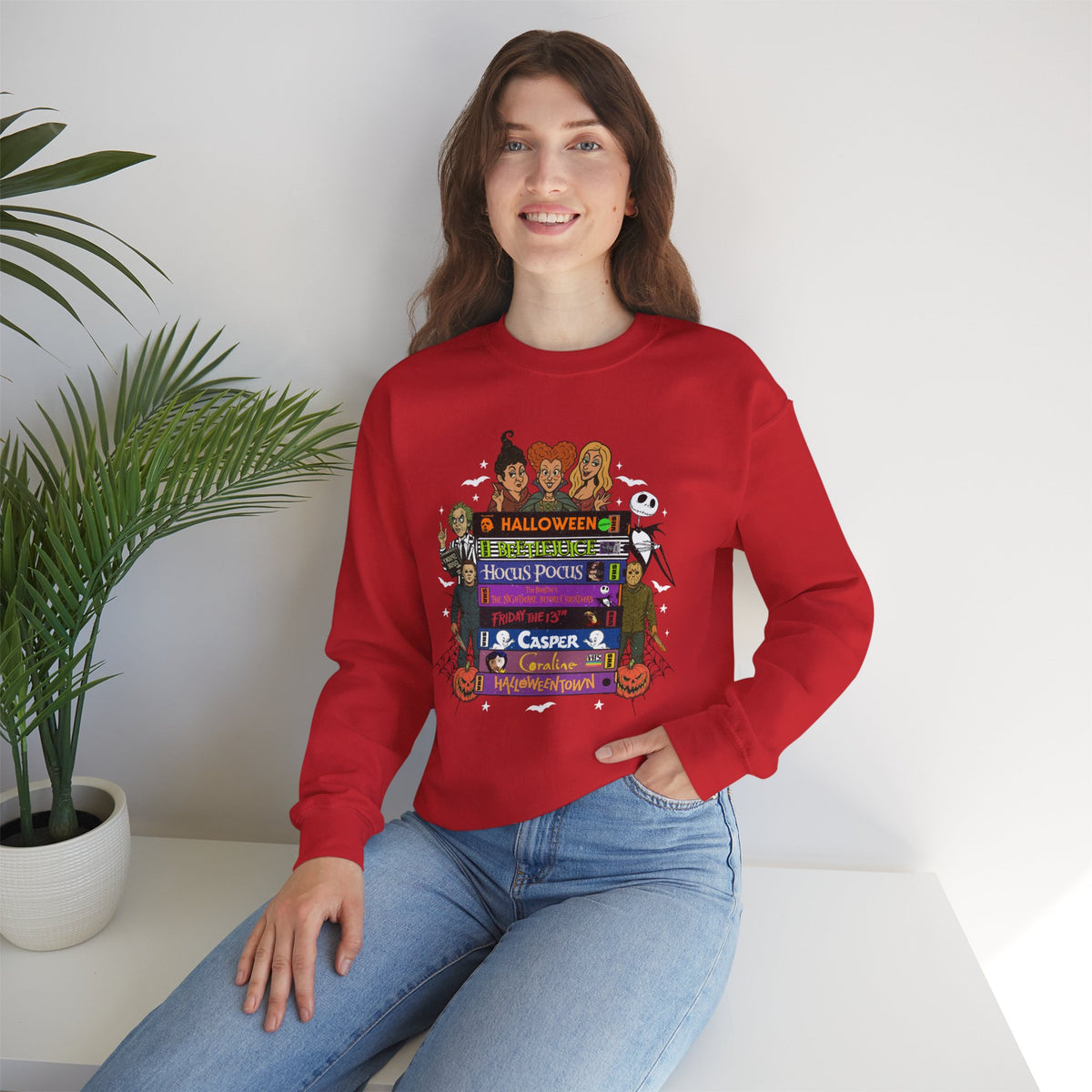 A woman wearing a red crewneck sweatshirt with a colorful Halloween-themed graphic print featuring VHS tapes, ghosts, and pumpkins, paired with blue jeans, sitting in a bright room next to a potted plant.