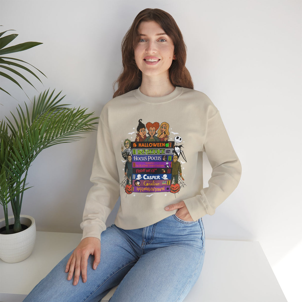 A woman wearing a beige crewneck sweatshirt with a colorful 'Spooky Movies' graphic on the front, paired with blue jeans, sitting in a bright room next to a potted plant.