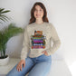 A woman wearing a beige crewneck sweatshirt with a colorful 'Spooky Movies' graphic on the front, paired with blue jeans, sitting in a bright room next to a potted plant.