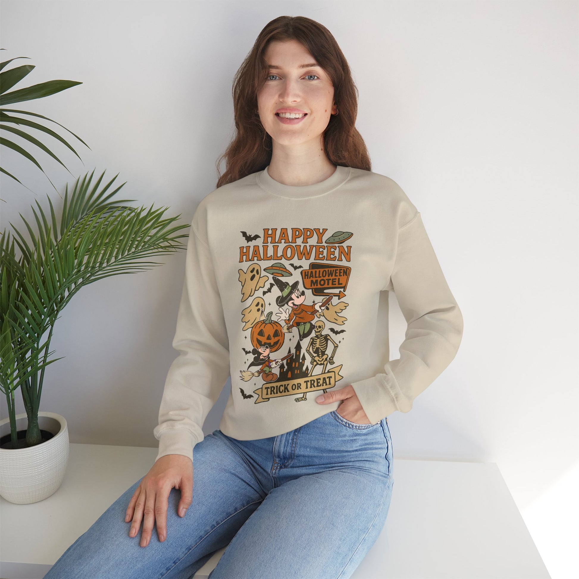 A woman wearing a beige crewneck sweatshirt with a colorful 'Happy Halloween' graphic print, paired with blue jeans, sitting on a white bench next to a potted plant.
