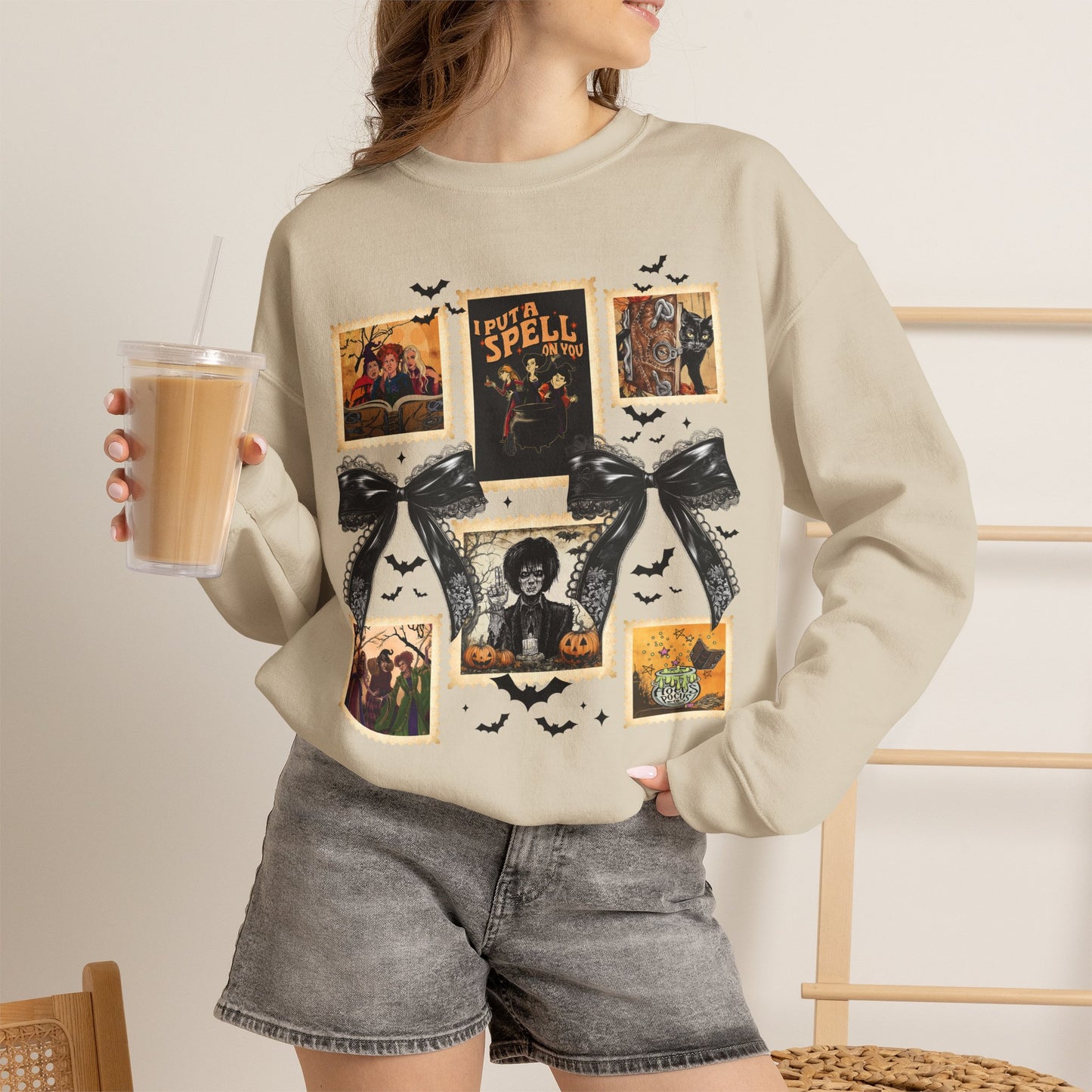 Woman wearing a beige crewneck sweatshirt featuring a vintage witch-themed Halloween collage with black bows, bats, pumpkins, and orange accents.
