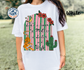 White t-shirt with a colorful graphic design featuring a cactus, tiger, and text, worn by a person outdoors.