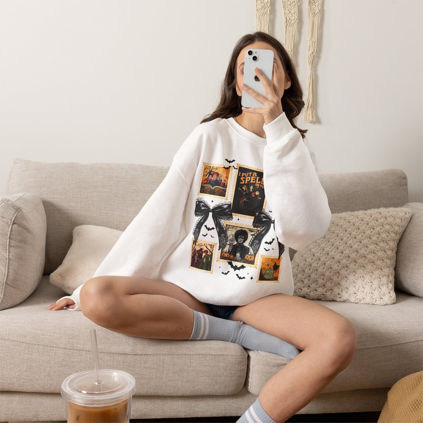 Woman wearing a white crewneck sweatshirt featuring a vintage witch-themed Halloween collage with black bows, bats, pumpkins, and orange accents.