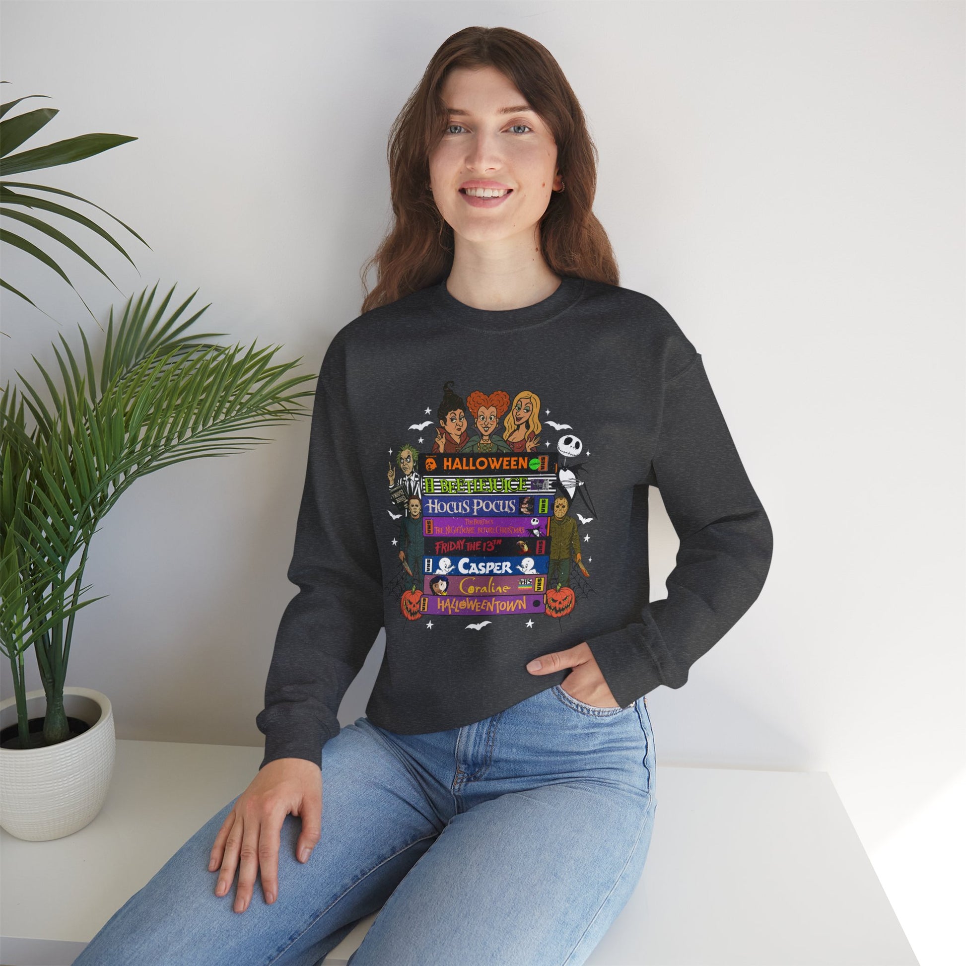 A woman wearing a dark gray crewneck sweatshirt with a colorful 'VHS tapes' graphic print, paired with blue jeans, sitting in a brightly lit room next to a potted plant.