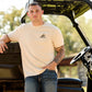 Ariat Mens Short Sleeve Shirt in beige cotton with a black Ariat logo and graphic on the chest, worn with blue jeans.