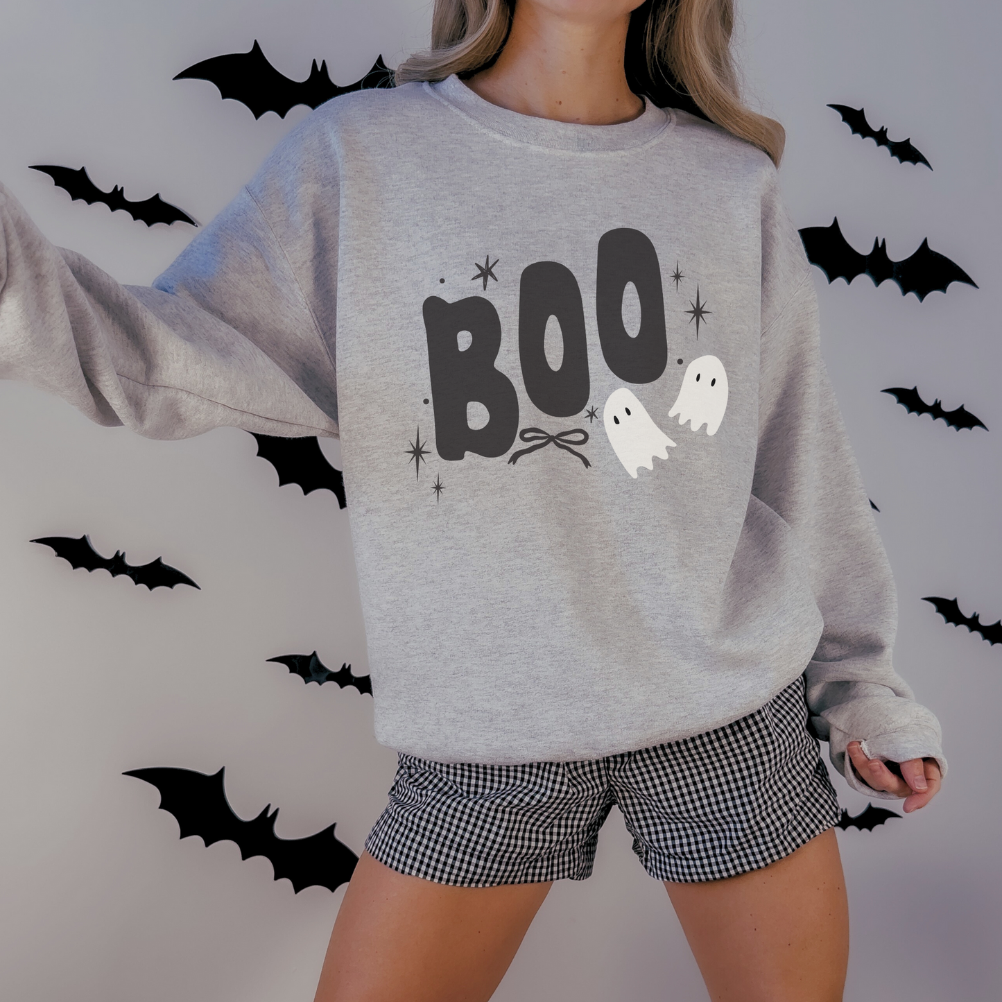 A woman wearing a light heather gray crewneck sweatshirt with a 'BOO' Halloween graphic, paired with black and white gingham shorts, standing against a wall decorated with black bat cutouts.