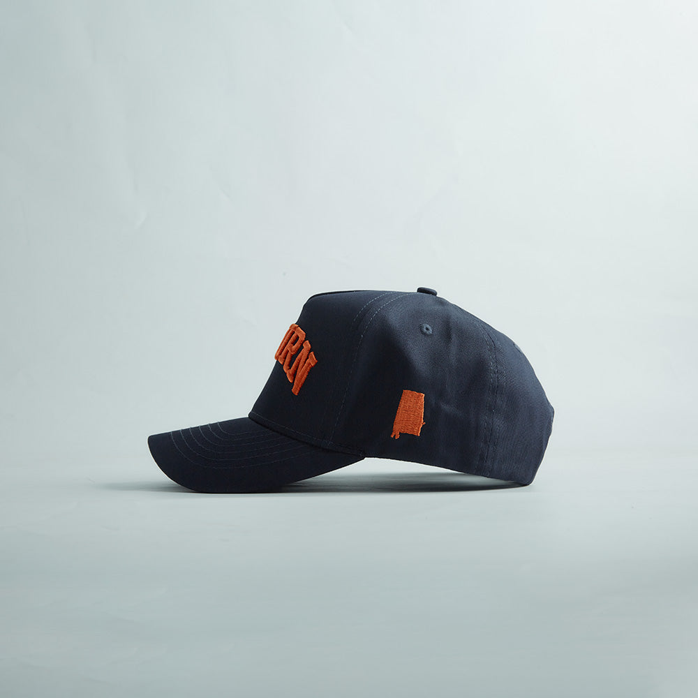 Navy blue snapback cap with curved bill, orange 3D "AUBURN" embroidery, and Alabama state silhouette, part of Auburn Alabama Legacy collection.