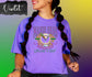 Person wearing a purple Mardi Gras t-shirt with colorful graphics and text on a gradient background.