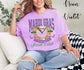Person wearing a purple 'Mardi Gras Social Club' t-shirt sitting on a couch.