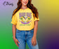 A woman wearing a bright yellow graphic t-shirt with 'Mardi Gras Social Club' printed on the front, paired with blue jeans, set against a vibrant green-to-yellow-to-purple gradient background with a 'Citrus' logo in the top left corner.