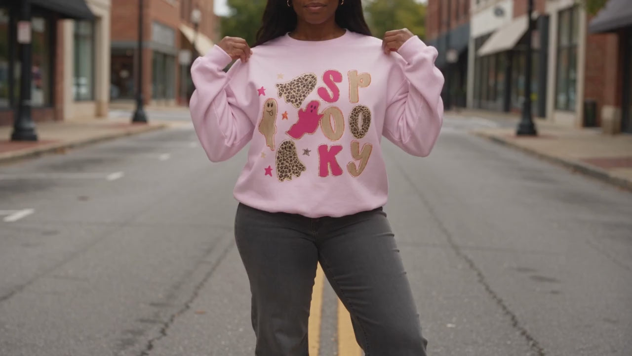 A woman modeling a light pink crewneck sweatshirt with a 'spooky graphic featuring cartoon ghost and stars