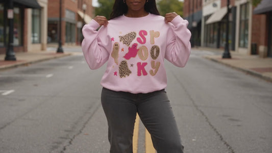 A woman modeling a light pink crewneck sweatshirt with a 'spooky graphic featuring cartoon ghost and stars