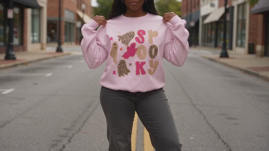 A woman modeling a light pink crewneck sweatshirt with a 'spooky graphic featuring cartoon ghost and stars