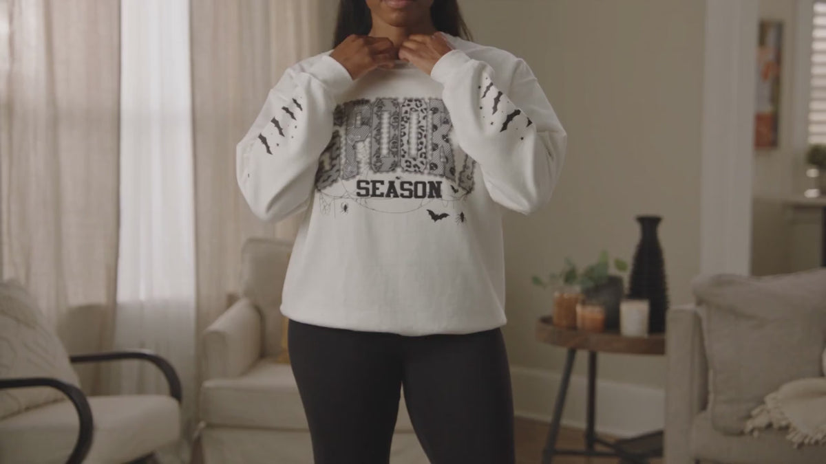A woman modeling while wearing a white crewneck sweatshirt with a distressed black graphic that reads 'spooky season'  in a patterned block.