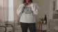A woman modeling while wearing a white crewneck sweatshirt with a distressed black graphic that reads 'spooky season'  in a patterned block.