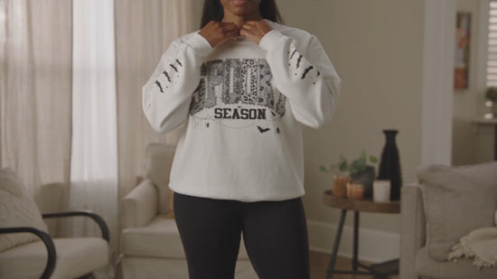 A woman modeling while wearing a white crewneck sweatshirt with a distressed black graphic that reads 'spooky season'  in a patterned block.