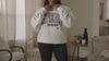 A woman modeling while wearing a white crewneck sweatshirt with a distressed black graphic that reads 'spooky season'  in a patterned block.