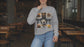 Woman modeling a Sport Grey crewneck sweatshirt featuring a vintage witch-themed Halloween collage with black bows, bats, pumpkins, and orange accents.
