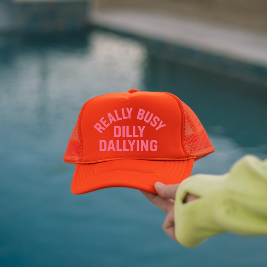 Really Busy Dilly Dallying Trucker Hat in bright orange foam with pink text, mesh sides, and curved brim