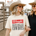Wranglin’ Blue Collar Cowboys Graphic Tee in cream cotton with red and black vintage western text design worn by a woman in a tan cowboy hat and sunglasses